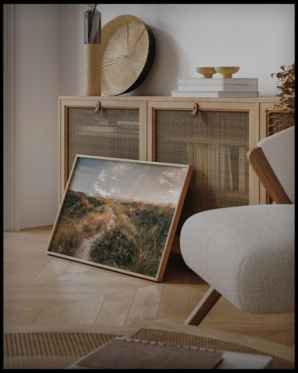 Ein gerahmtes Poster von einer Wanderung durch grasige Dünen, an einer Holzkommode angelehnt und stehend auf dem Holzboden