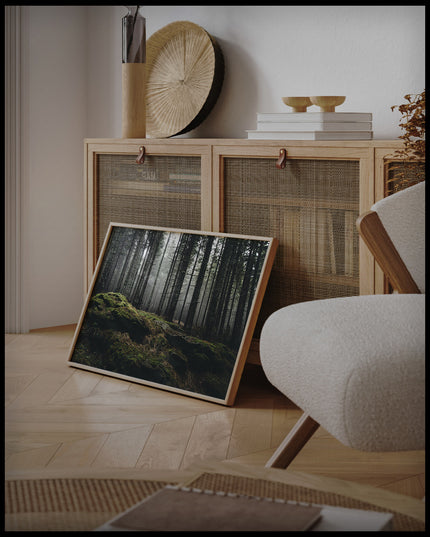 Ein gerahmtes Poster von einem nebeligen Wald mit moosbedeckten Steinen im Vordergrund, an einer Holzkommode angelehnt und stehend auf dem Holzboden