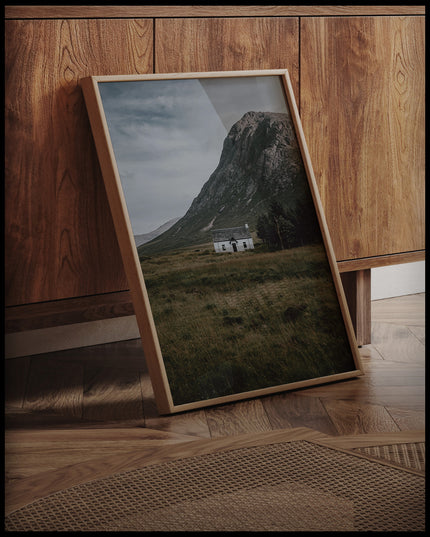 Ein gerahmtes Poster von einer weißen Holzhütte auf einer Wiese vor einem Berg, an einer Holzkommode angelehnt und stehend auf dem Holzboden