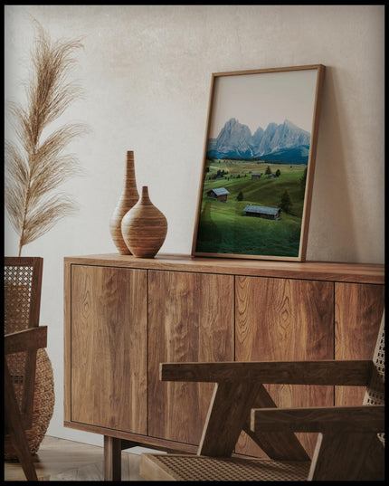 Ein gerahmtes Poster von einer grünen Alm mit kleinen Hütten und den Dolomiten im Hintergrund, auf einer Holzkommode an die Wand angelehnt