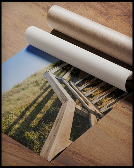 Ein Poster von einer Holztreppe die über Dünen am Strand führt, gerollt auf einer Holzoberfläche liegend