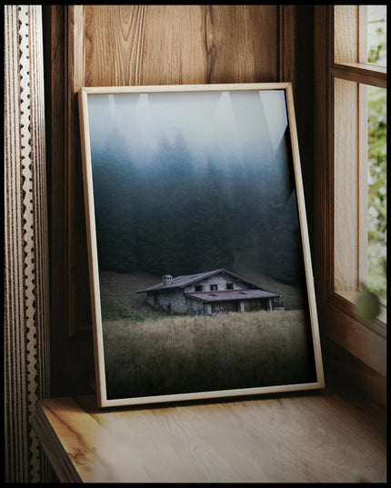 Ein gerahmtes Poster von einer einsamen Hütte im Nebel, umgeben von hohen Bäumen, vor einem Fenster an die Wand angelehnt