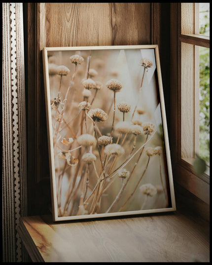 Ein gerahmtes Poster von vertrockneten Blumen in weichen Braun- und Beigetönen, vor einem Fenster an die Wand angelehnt