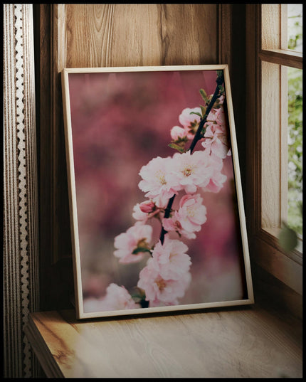 Ein gerahmtes Poster von einem Zweig mit zarten, rosa Kirschblüten vor einem unscharfen Hintergrund, vor einem Fenster an die Wand angelehnt