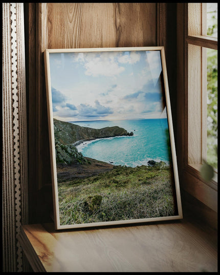 Ein gerahmtes Poster vom Blick einer Bucht über das blaue Meer, vor einem Fenster an die Wand angelehnt