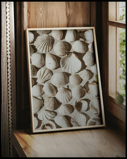 Ein gerahmtes Poster von mehreren weißen Muscheln auf beigefarbenen Untergrund, vor einem Fenster an die Wand angelehnt