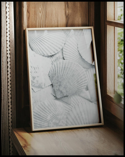 Ein gerahmtes Poster von mehreren weißen Muschelschalen, die dicht beieinander liegen und eine strukturierte Oberfläche bilden, vor einem Fenster an die Wand angelehnt