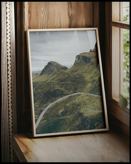 Ein gerahmtes Poster von einer Straße die an mehreren bewachsenen, kargen Bergen vorbei führt, vor einem Fenster an die Wand angelehnt