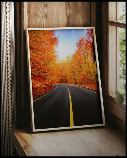 Ein gerahmtes Poster von einer leeren schwarzen Straße die durch einen herbstlichen. orangenen Wald führt, vor einem Fenster an die Wand angelehnt