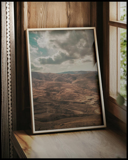 Ein gerahmtes Poster von einer trockenen, hügeligen Landschaft, vor einem Fenster an die Wand angelehnt