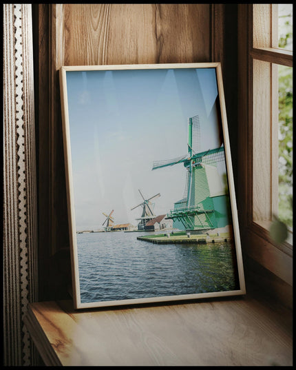 Ein gerahmtes Poster von drei Windmühlen aus Holz die am Wasser stehen, vor einem Fenster an die Wand angelehnt